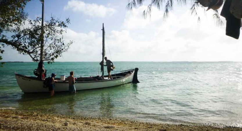 a sailboat floats in shallow blue water on an outward bound expedition. A couple of people are standing in the knee-deep water, and a couple are standing in the boat. 